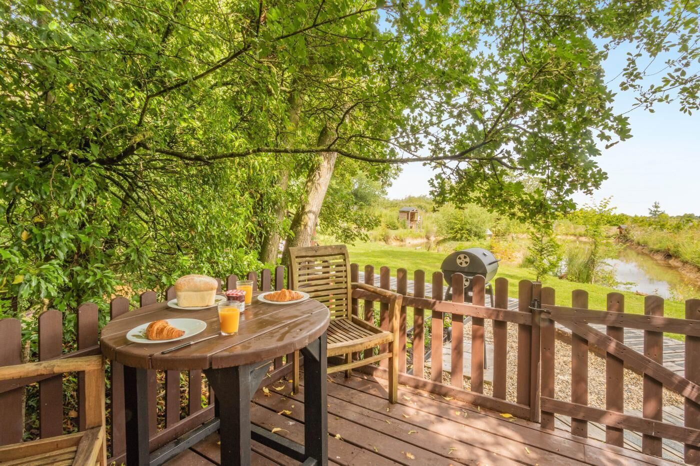 Ein Essbereich im Freien mit einer Holzterrasse, einem Tisch mit Speisen und Getränken, einem Grill und Blick auf einen Teich und Grünflächen.