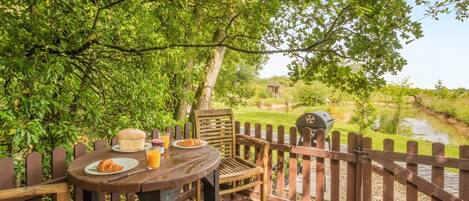 Ein Essbereich im Freien mit einer Holzterrasse, einem Tisch mit Speisen und Getränken, einem Grill und Blick auf einen Teich und Grünflächen.