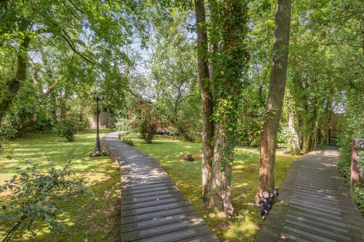 A wooden pathway in a lush garden with trees and a wooden structure in the background.