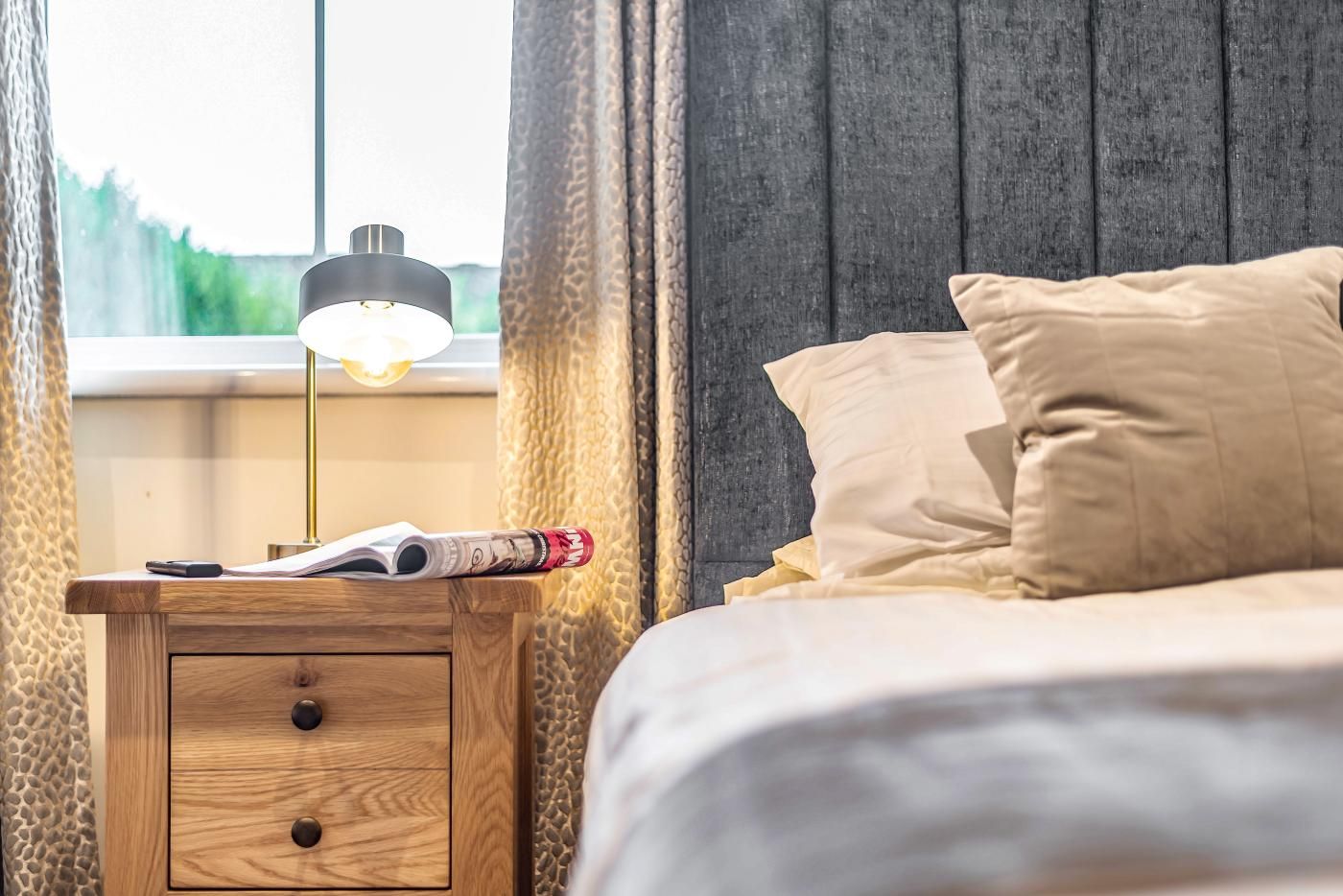 A neatly made bed with a headboard, a bedside table with a lamp, and a window with curtains.