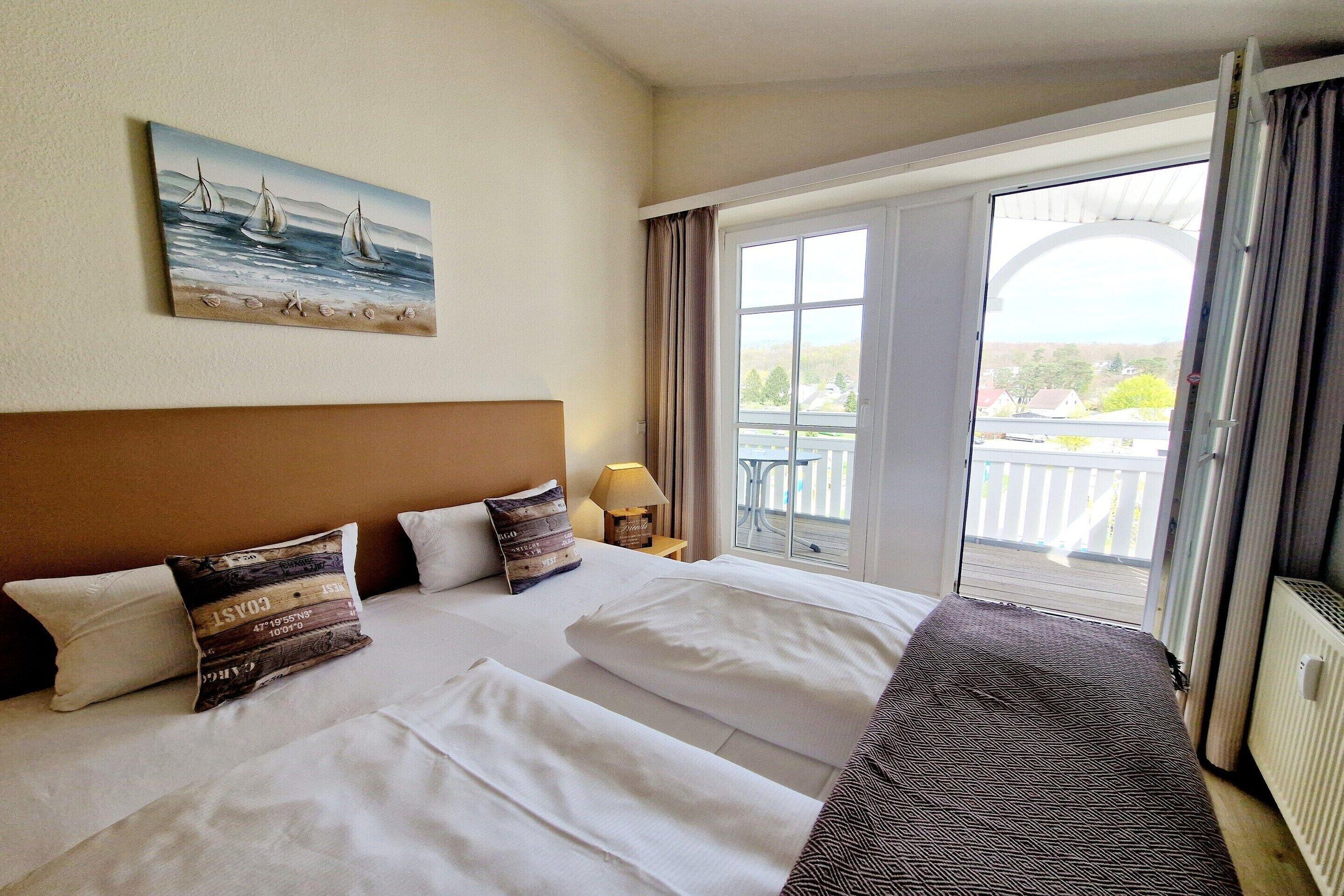 Quarto de hotel com duas camas, um quadro de veleiros, uma varanda com vista e uma janela com cortinas.