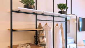 A modern room with a metal shelving unit, a safe, a small table with a handbag and shoes, and a bed with a white headboard. - Urban Residence: Stylish comfort with city views in the heart of Piraeus. (Pireas)