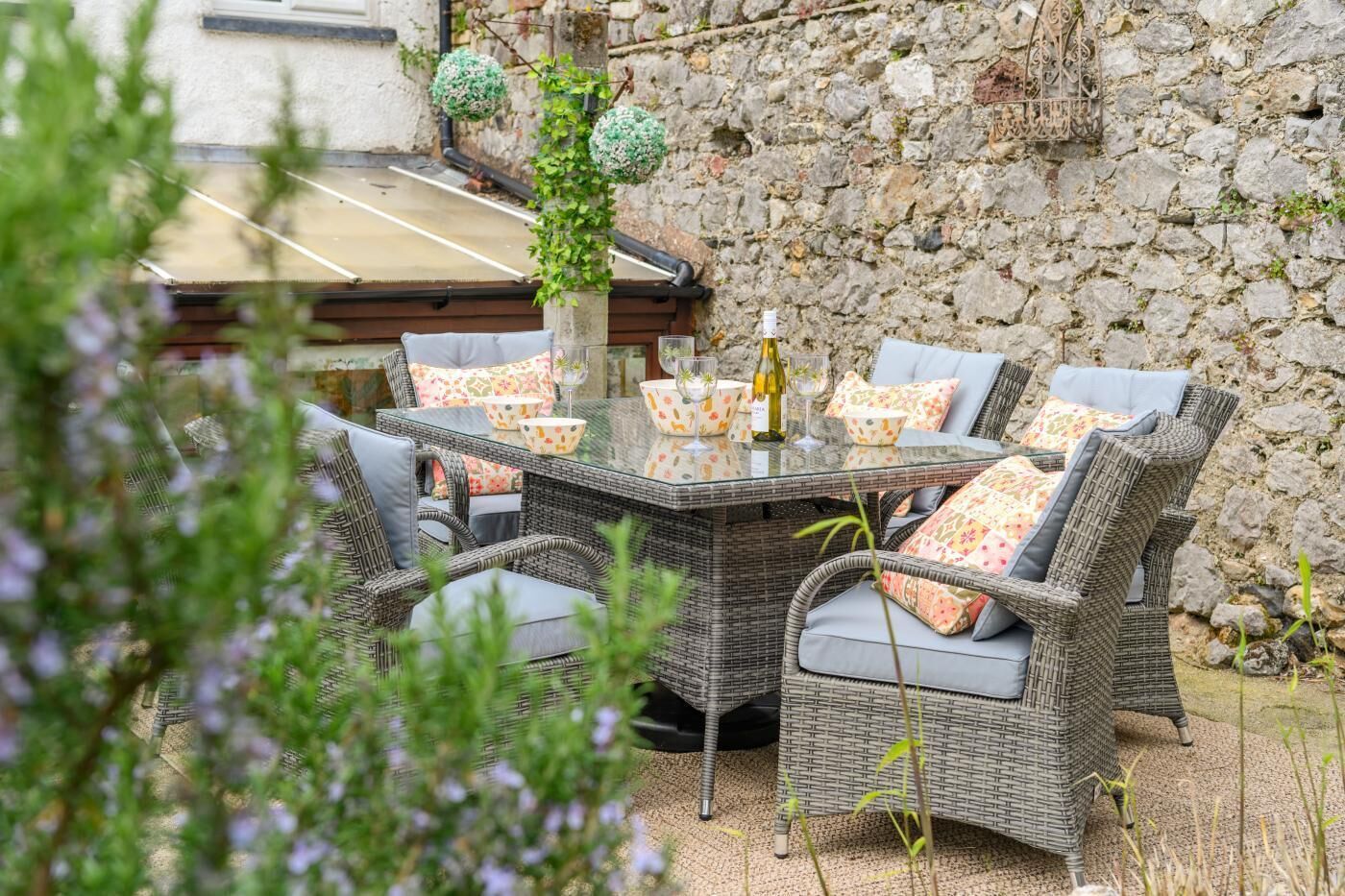 An outdoor dining area with wicker furniture and a glass table, set against a stone wall.