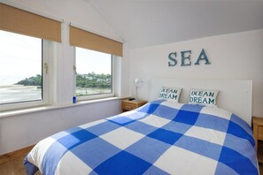 Un dormitorio con colcha a cuadros, dos almohadas con el texto 'OCEAN DREAM', una ventana con vistas al mar y un letrero con la palabra 'SEA' en la pared.