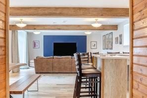 A modern living room with a sofa, a wooden bench, a dining area with bar stools, and a large TV screen. - 8 Bed in Buxton (oc-w34369) (Hartington Nether Quarter)