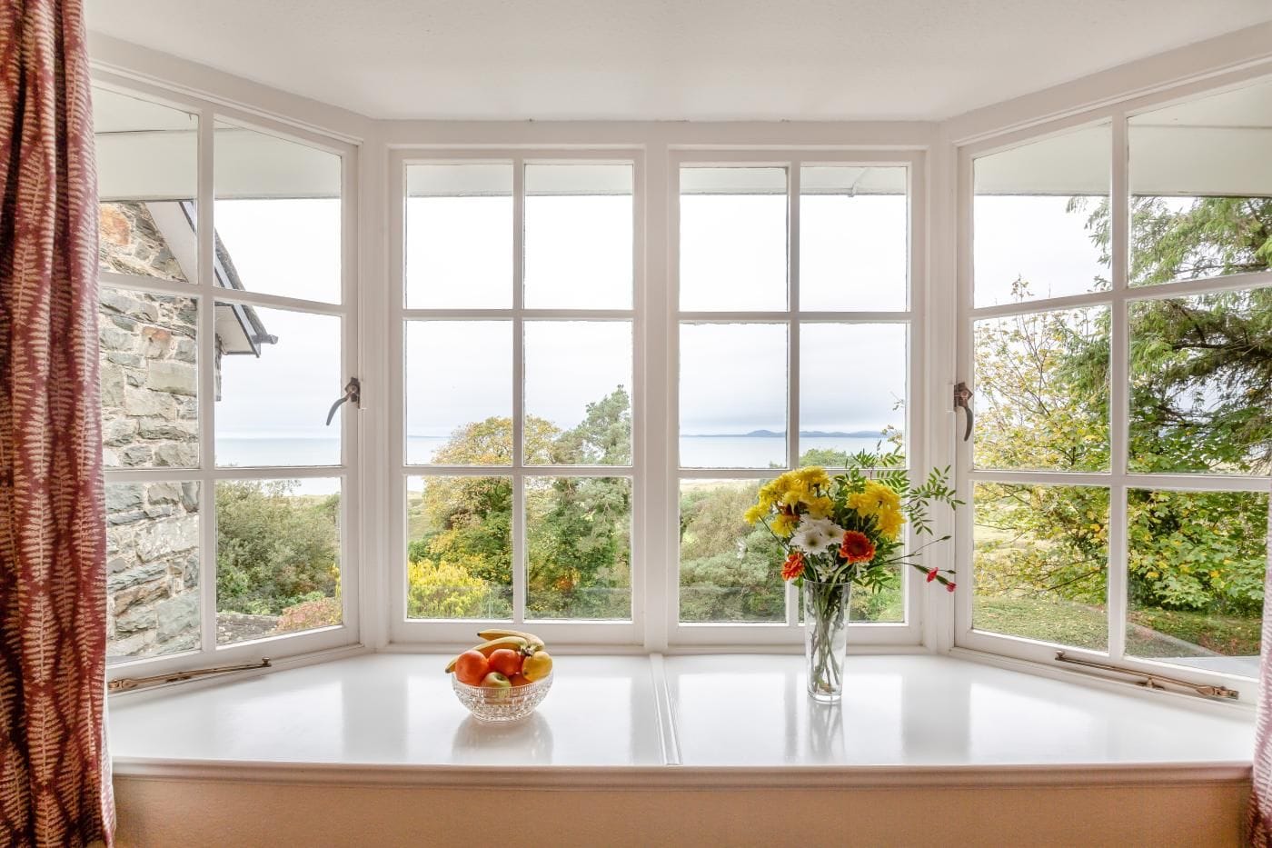 A bay window with a clear view of the outdoors, a bowl of fruit on the windowsill, and a vase with flowers.