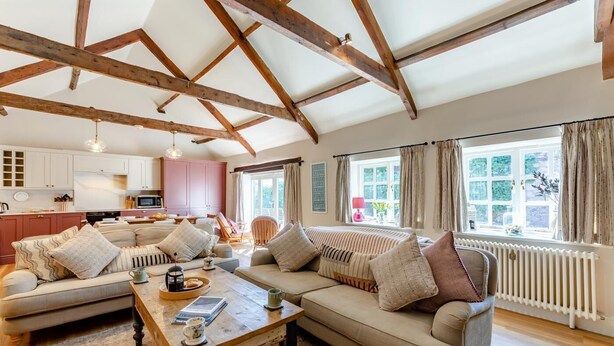 A spacious living room with a high ceiling and exposed wooden beams, a cozy sofa set, a wooden coffee table, and a view of the kitchen and garden through large windows. - 3 Bed in Bamburgh (oc-t34385) (Bamburgh)