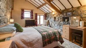 Une chambre confortable avec un mur en pierre, un plafond en bois, un lit recouvert d’une couvre-lit à motifs floraux, un bureau avec une lampe et une cheminée en pierre.