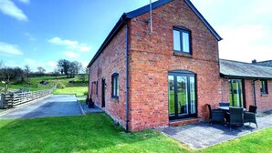 A brick building with a large glass door, a small patio with outdoor seating, and a grassy area with a fence. - 2 Bed in Llandinam (oc-waa377) (Llandinam)