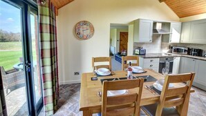 A dining area with a wooden table and chairs, a clock on the wall, and a view of the outdoors through a glass door. - 2 Bed in Llandinam (oc-waa377) (Llandinam)