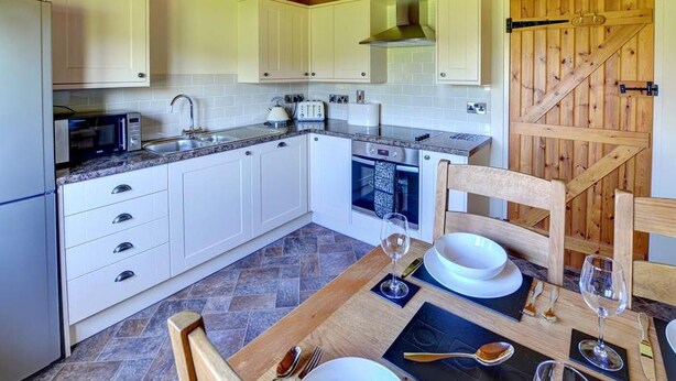 A kitchen with white cabinets, a microwave, a toaster, and a wooden dining table set with plates and wine glasses. - 1 Bed in Llandinam (oc-waa376) (Llandinam)