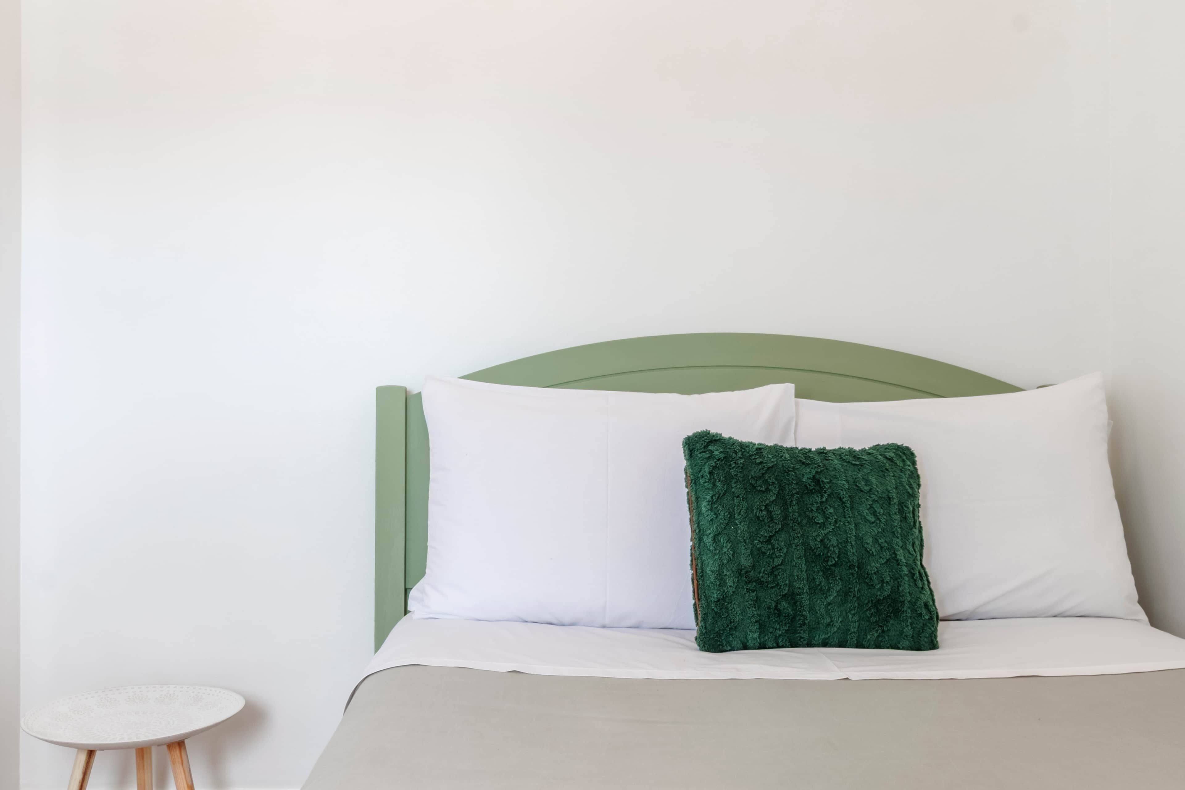 Ein Bett mit weißer Bettwäsche und einem grünen Kopfteil, ein kleiner runder Tisch mit weißer Tischplatte und Holzbeinen sowie ein grünes Zierkissen.