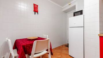 Uma cozinha compacta com paredes revestidas de azulejos brancos, uma geladeira branca, uma mesa de jantar com toalha vermelha e cadeiras brancas.