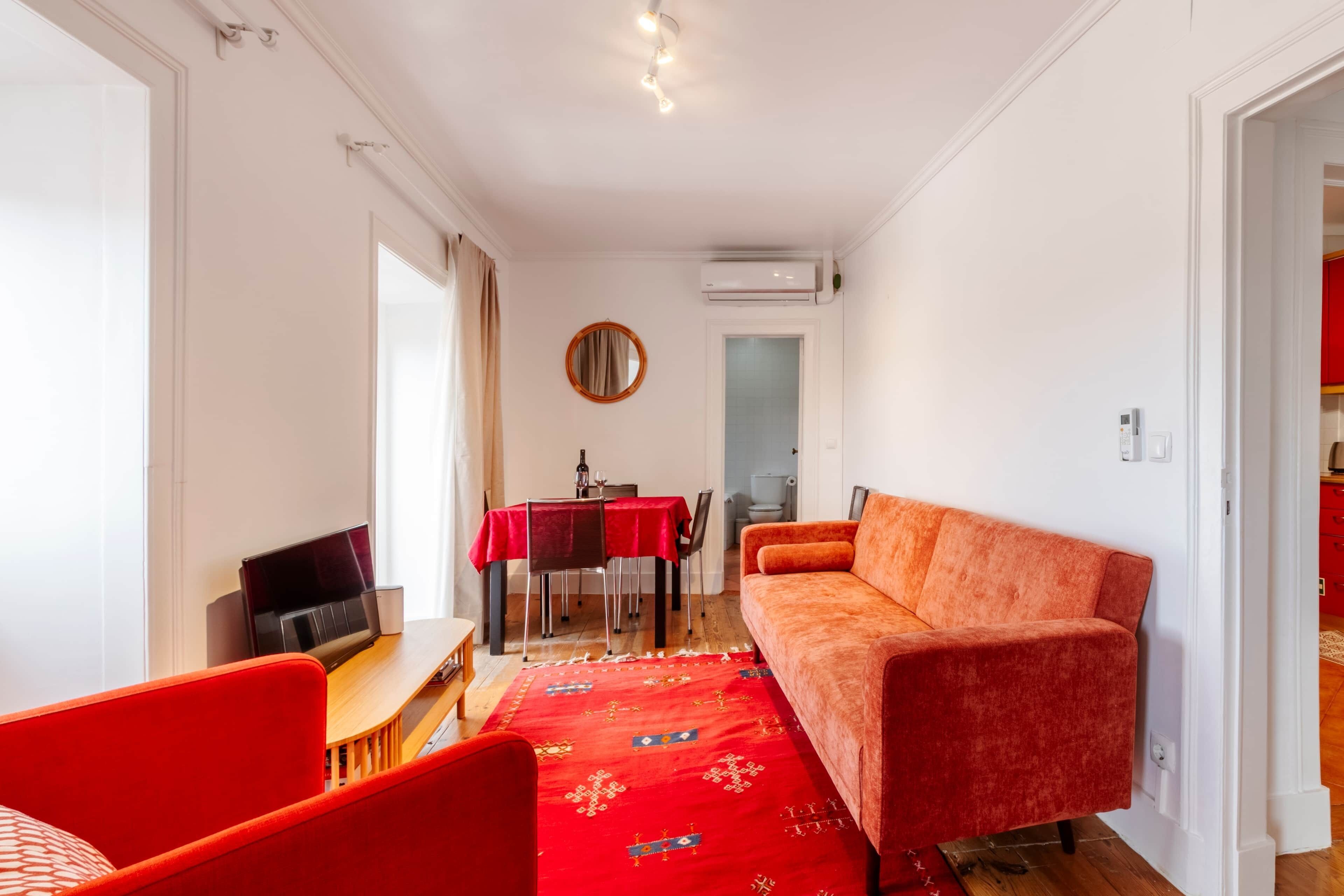 A room with a red sofa, a TV, a dining table with chairs, and a red carpet.
