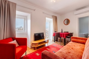 A living room with a red sofa, a TV on a wooden stand, a dining area with a table and chairs, and large windows with curtains. - Panorama View Apartment in Amazing Viewpoint (Lisbon)