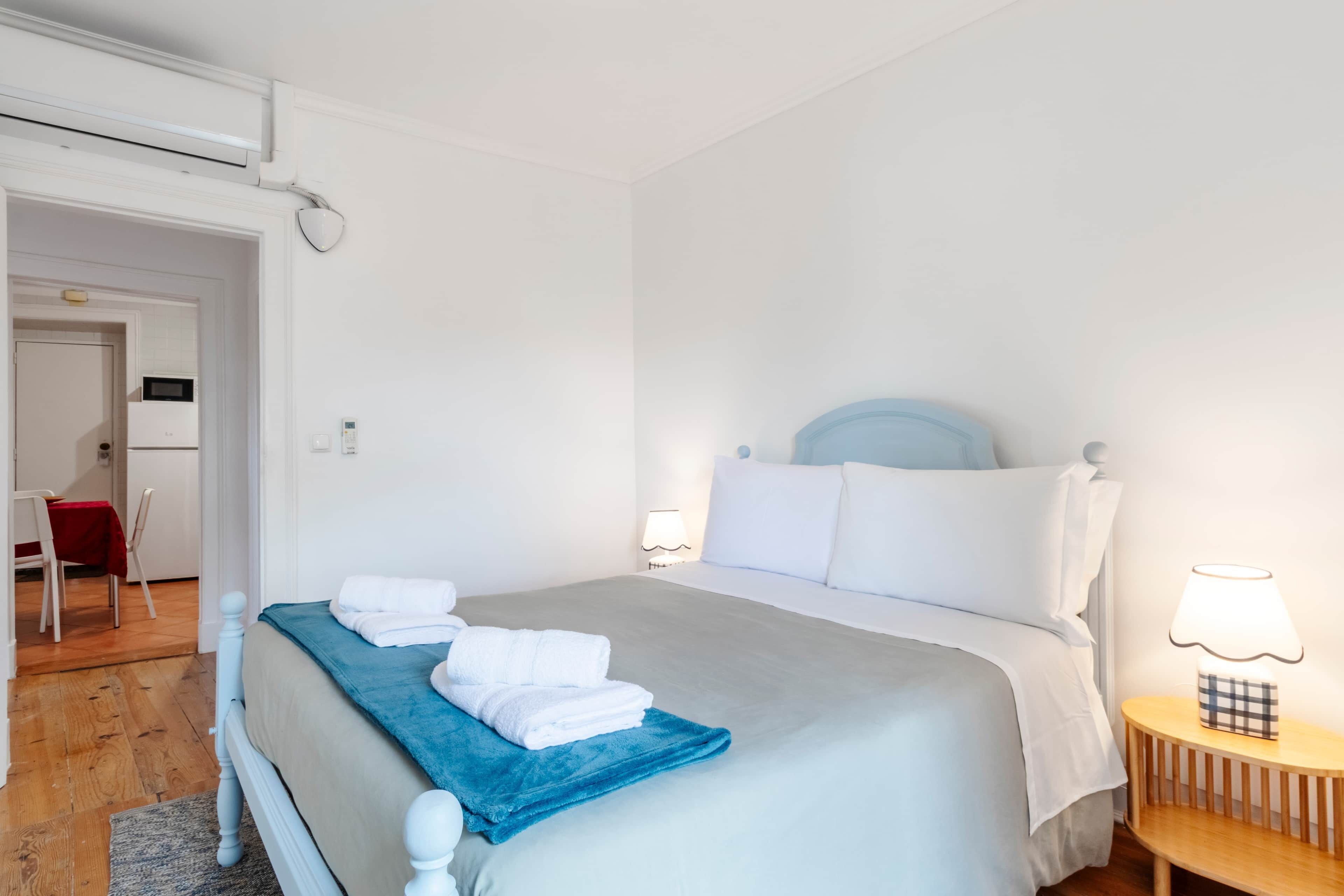 A hotel room with a bed, a bedside table with a lamp, a chair, and a desk with a red tablecloth.
