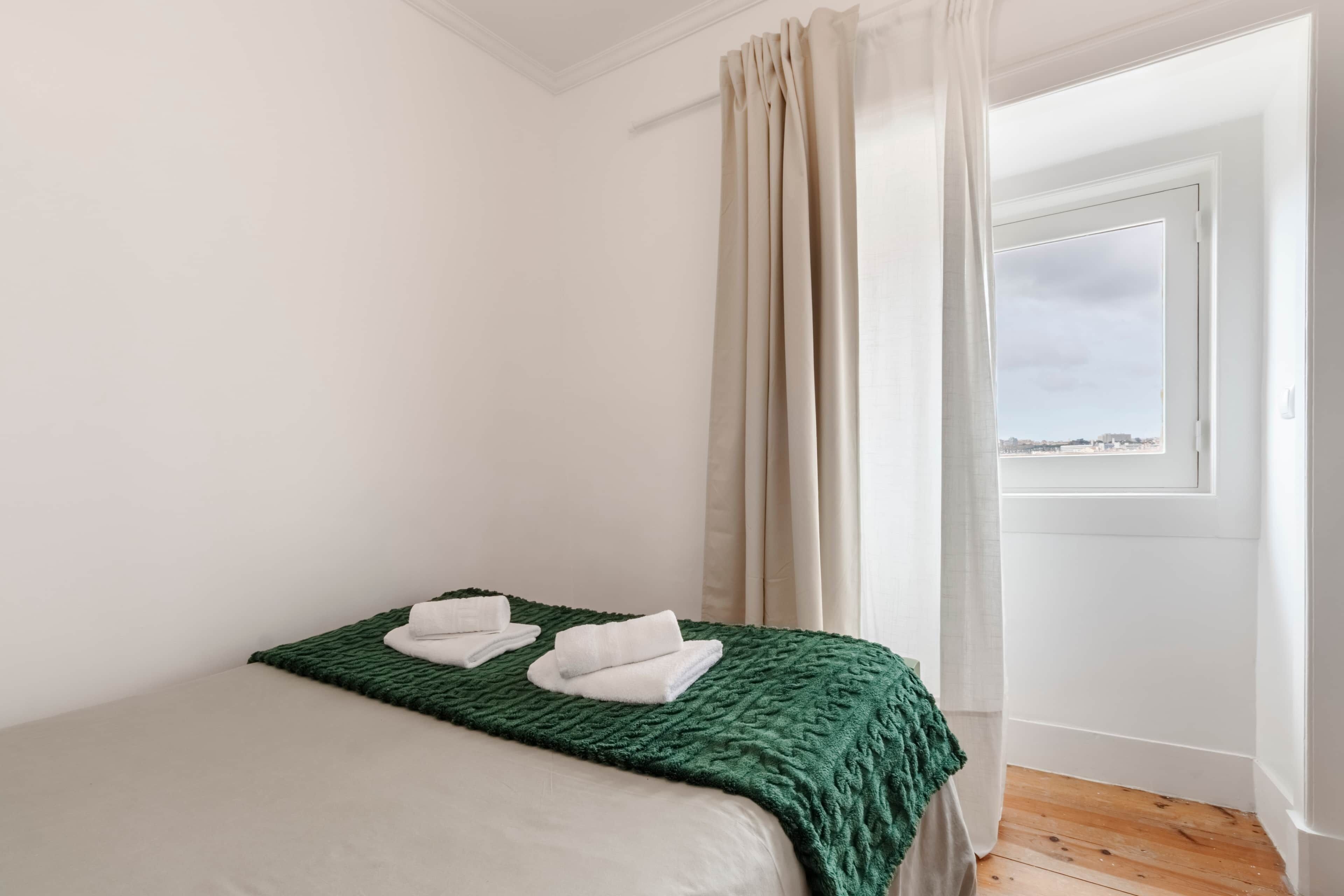 A bedroom with a bed, green blanket, white towels, a window with curtains, and a view of the city.