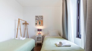 Et hotelværelse med to senge, et lille bord med lampe, et billede på væggen og et vindue med gardiner.