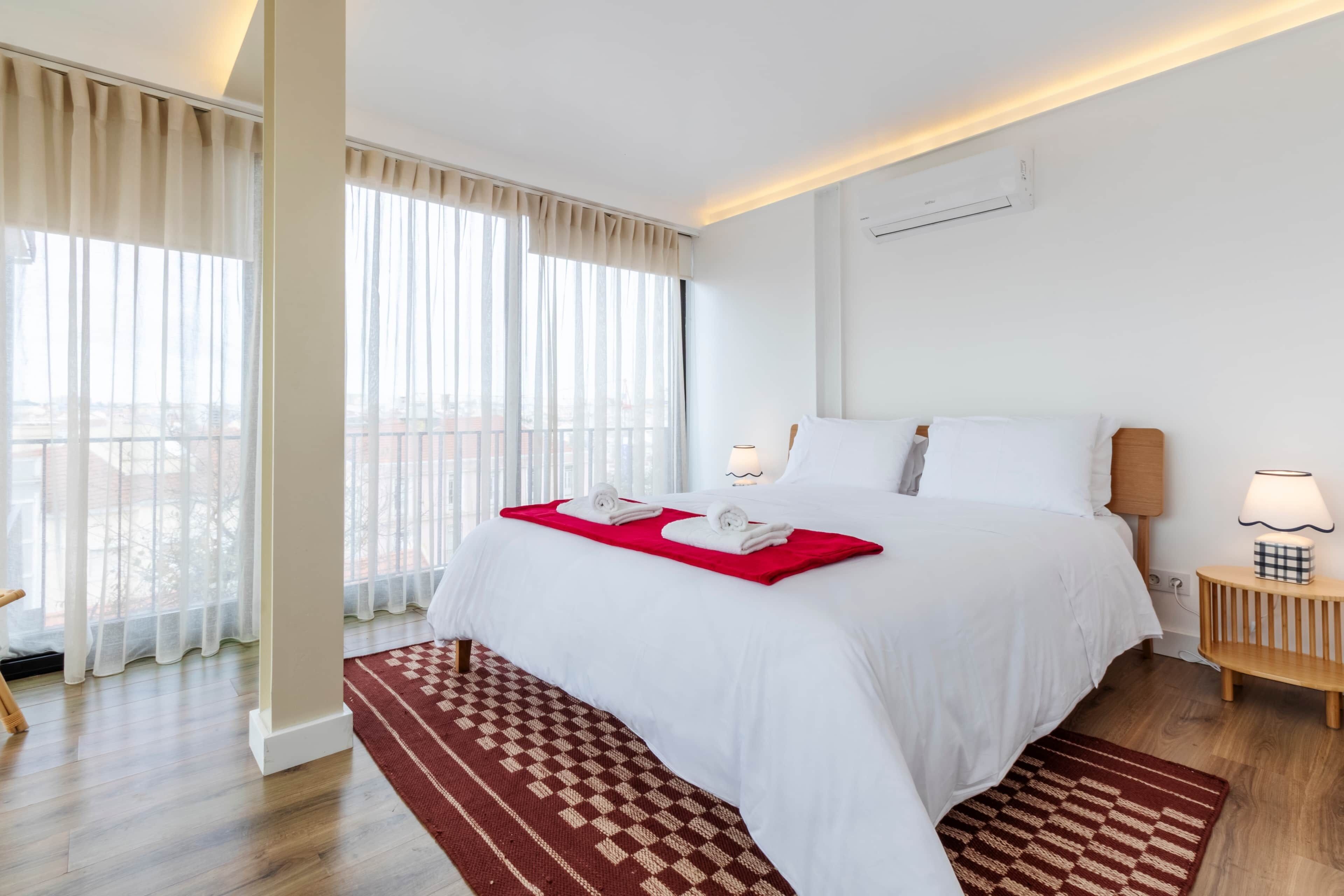 A hotel room with a large bed, a bedside table with a lamp, a window with sheer curtains, and a view of the city.