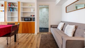 Ett modernt vardagsrum med en soffa, ett matbord med stolar, en bokhylla, en köksdel och en stor spegel på väggen.