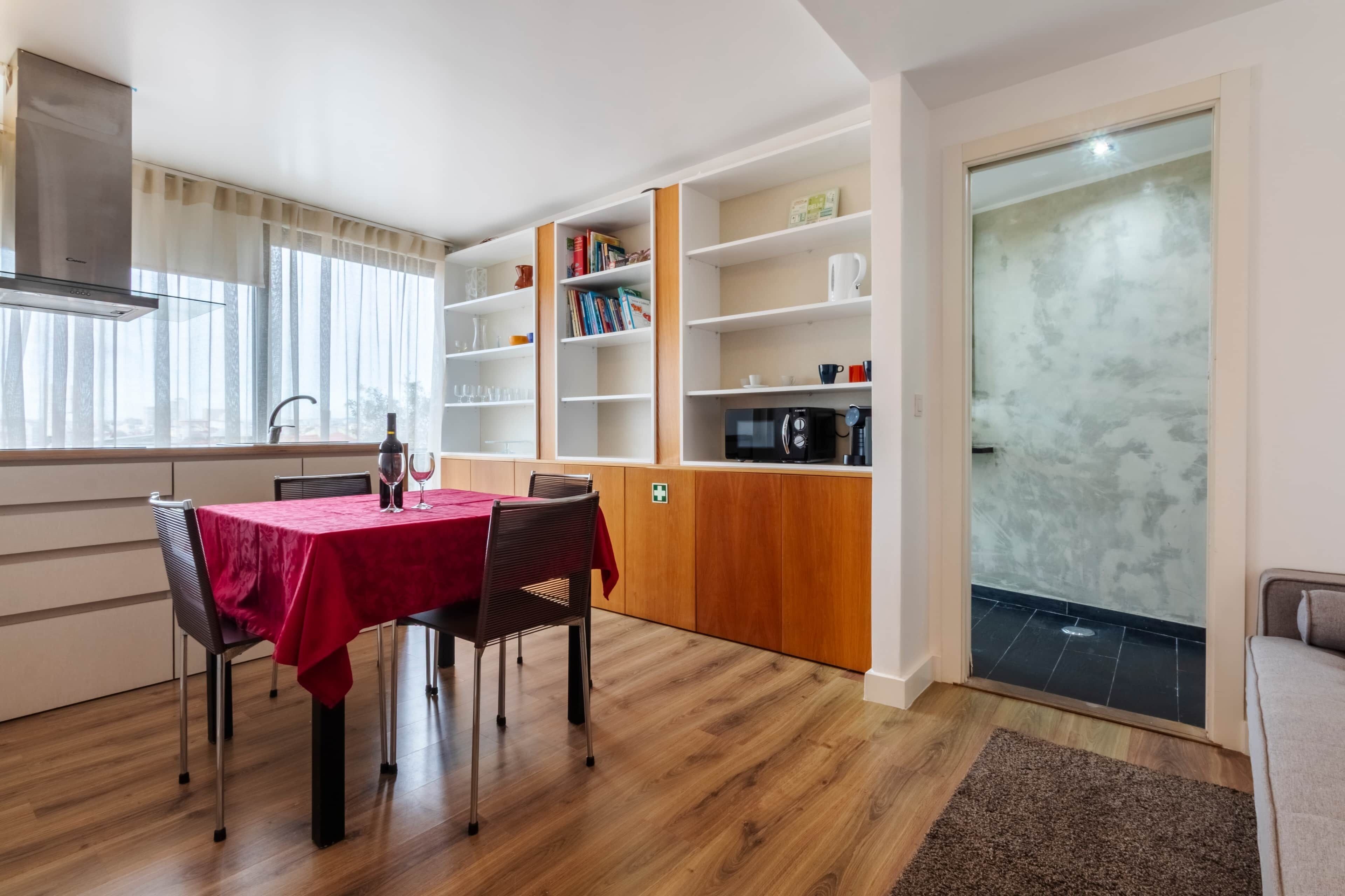 Een eetgedeelte met een tafel, stoelen en een rode tafelkleed. Er staan boekenkasten met boeken en decoratieve objecten. Een magnetron is zichtbaar op het aanrecht.
