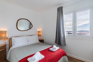 A hotel room with a bed, a mirror, two bedside lamps, a window with curtains, and a view of the cityscape. - Panorama View Apartment in Beautiful Viewpoint (Lisbon)