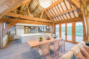 Un interior espacioso con una gran mesa de madera para comer, sillas, una zona de cocina y vistas a través de puertas de cristal amplias.