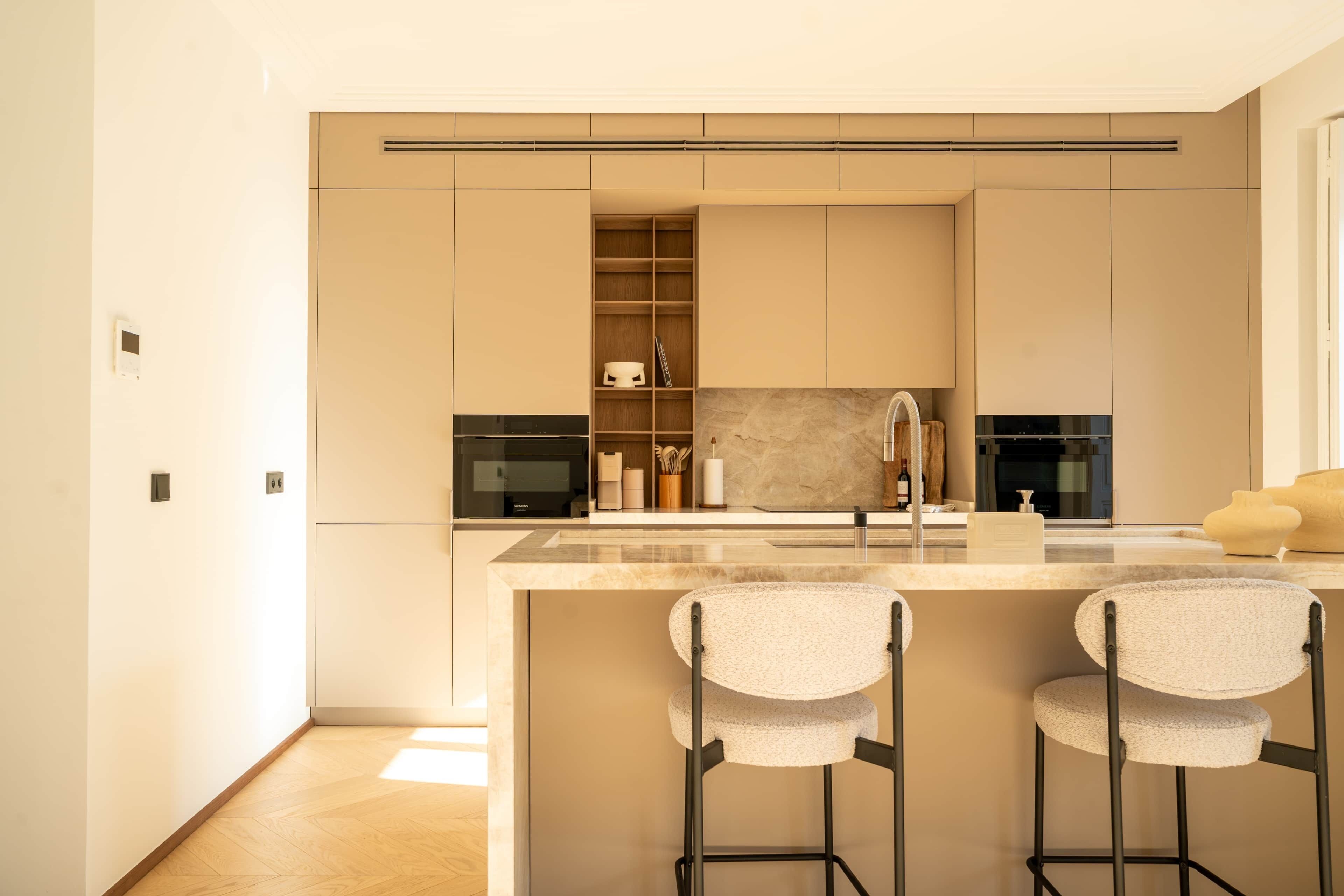 Una cocina moderna con encimera de mármol, dos taburetes de bar, electrodomésticos integrados y una estantería de madera.