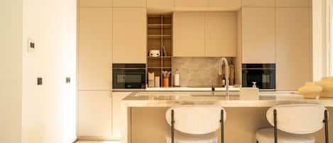 Una cocina moderna con encimera de mármol, dos taburetes de bar, electrodomésticos integrados y una estantería de madera.