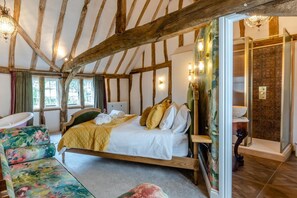 A rustic bedroom with exposed wooden beams, a bed with white linens, a floral sofa, and a shower area with a decorative wall. - 11 Bed in Stebbing (oc-p34450) (Stebbing Green)