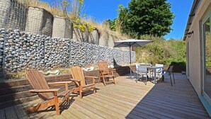 Una terraza de madera con mobiliario exterior, un muro de piedra y una puerta de cristal que da a un porche cubierto.