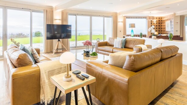 Ein großzügiger Wohnbereich mit Ledersofas, einem Flachbildfernseher und großen Fenstern mit Blick ins Freie.