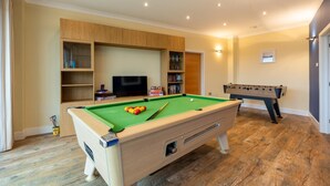 Une table de billard recouverte d’un drap vert, une table de baby-foot et un téléviseur dans une pièce dotée de meubles et d’étagères en bois.