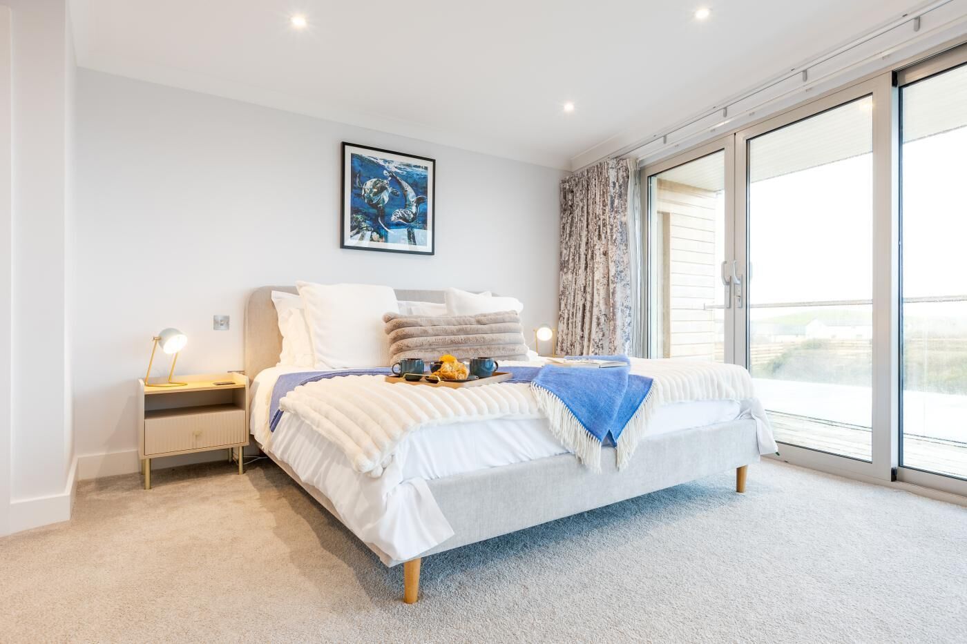 A modern bedroom with a bed, a nightstand, a desk, a window with a view, and a painting on the wall.