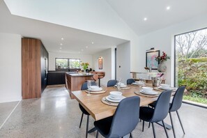 A modern dining area with a wooden table set for six, a kitchen in the background, and a large window with a view of greenery. - 3 Bed in Leominster (oc-w34380) (Leominster)