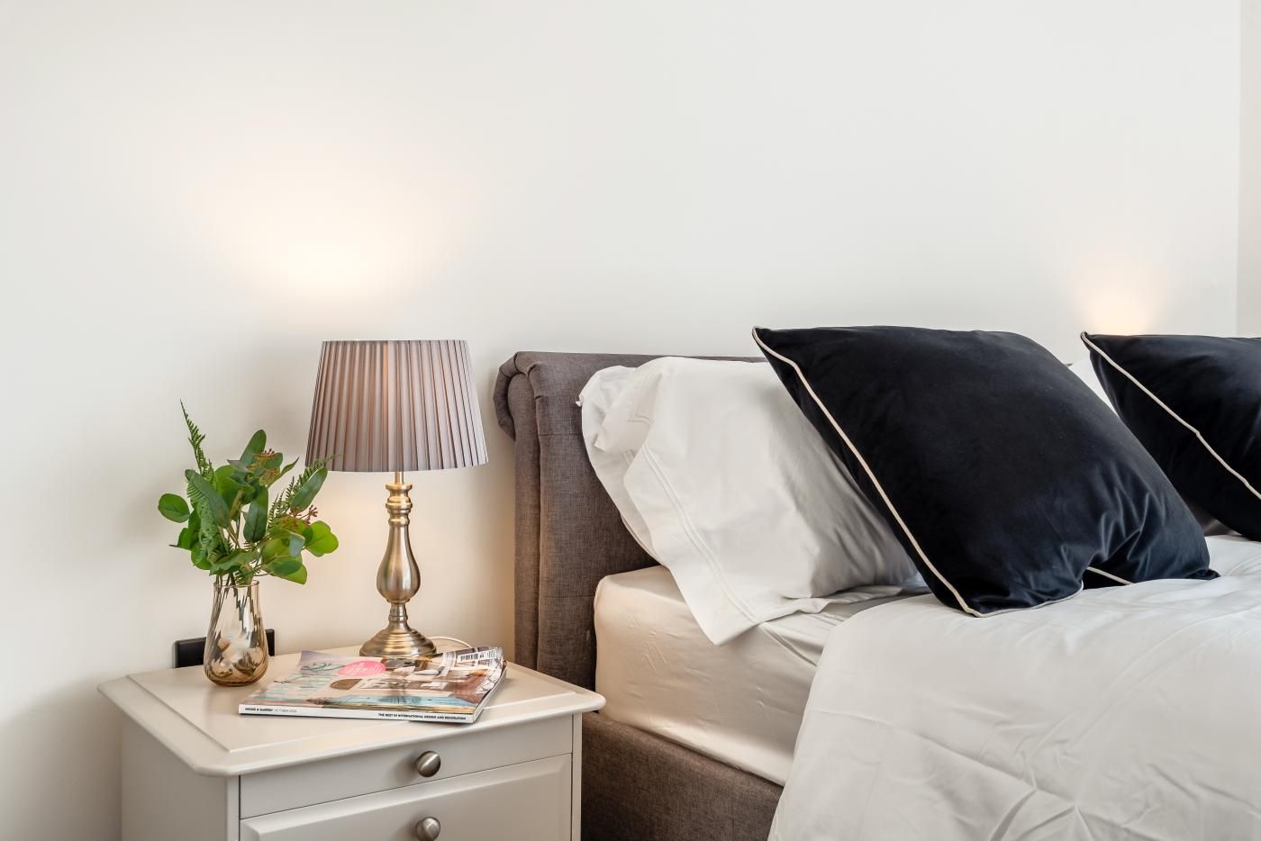 A neatly arranged bedroom with a bedside table, lamp, magazine, and a plant.