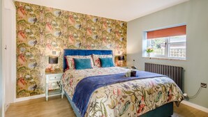 Une chambre à coucher avec un papier peint à motifs floraux, une tête de lit bleue en tissu, une table de chevet avec une lampe et une fenêtre donnant sur un beau paysage.