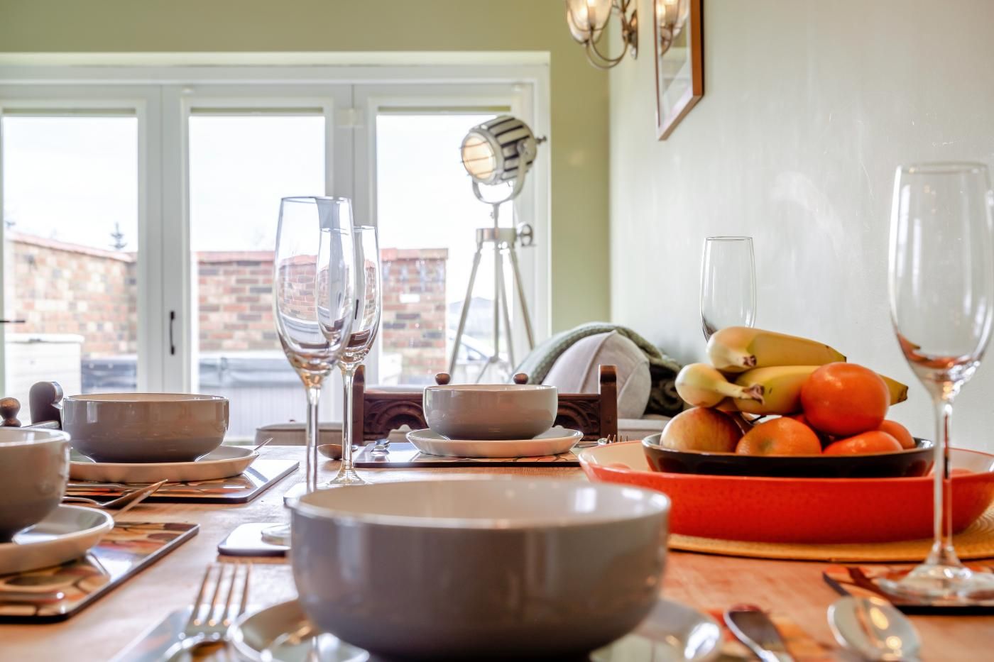 A dining table set with various bowls, plates, and cutlery, a fruit bowl with bananas and oranges, and two wine glasses.
