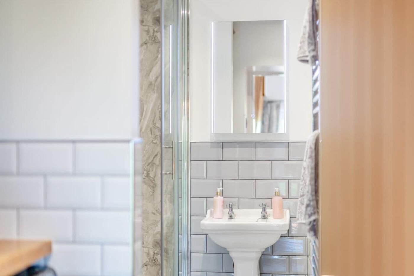 A bathroom with a marble wall, a glass shower enclosure, a white sink with two pink soap dispensers, and a mirror above the sink.