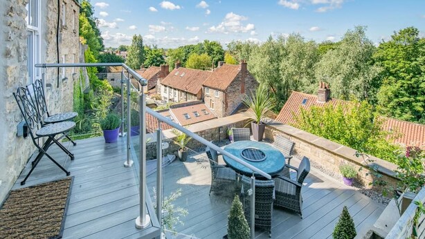 Eine Holzterrasse mit Gartenmöbeln, einem Metallgeländer und Blick auf umliegende Gebäude und Grünflächen.