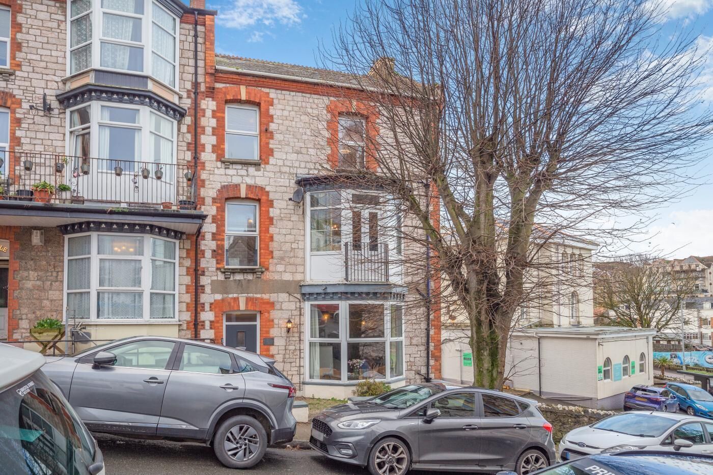 Une rangée de maisons de ville avec des bow-windows, un arbre au premier plan et des voitures garées.