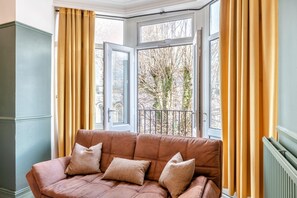 Une pièce avec un canapé en cuir marron, une grande fenêtre agrémentée de rideaux et une vue sur les arbres à l’extérieur.