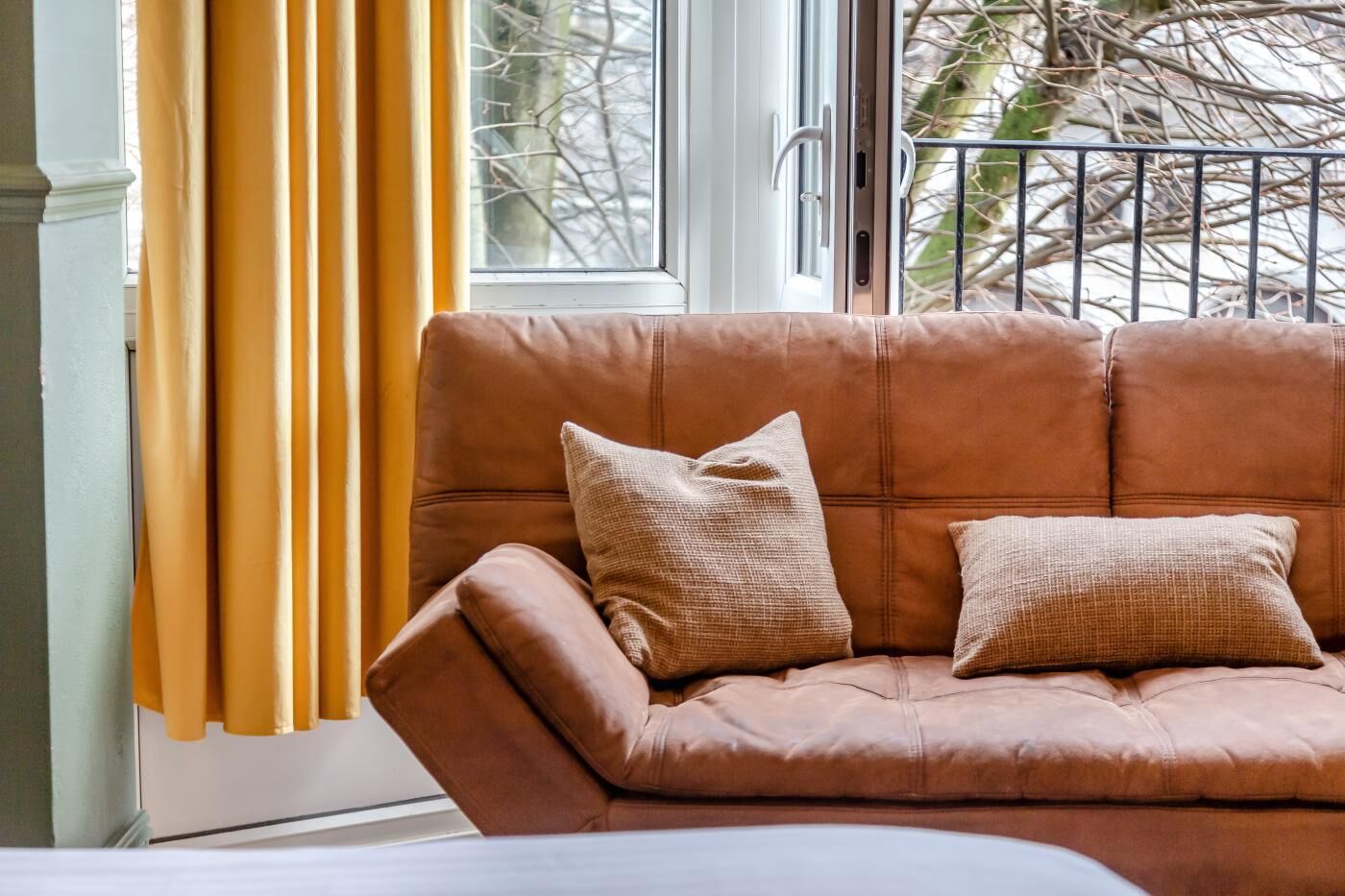 A brown leather sofa with two cushions, positioned next to a window with yellow curtains.