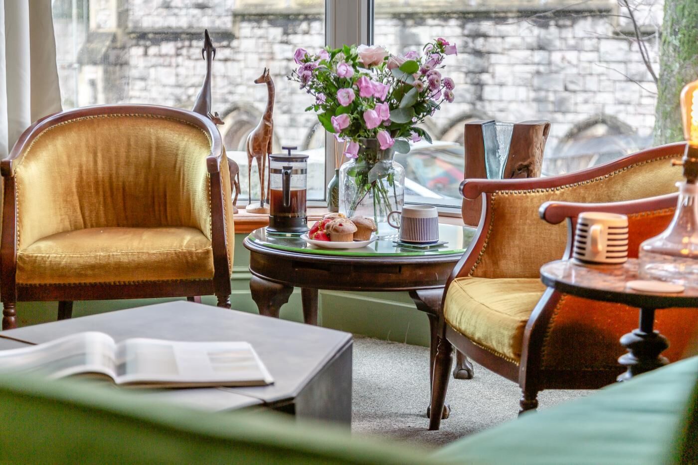 A room with two upholstered chairs, a round table with a vase of flowers, a book, and a window with a view of a stone building.