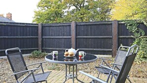 Et rundt bord med stole, en tepotte og to kopper på en terrasse med et træhegn i baggrunden.