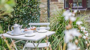A table with a pitcher, teacups, and a plate with pastries, set outdoors with greenery and a stone wall in the background. - 2 Bed in Padstow (oc-p00557) (Padstow)
