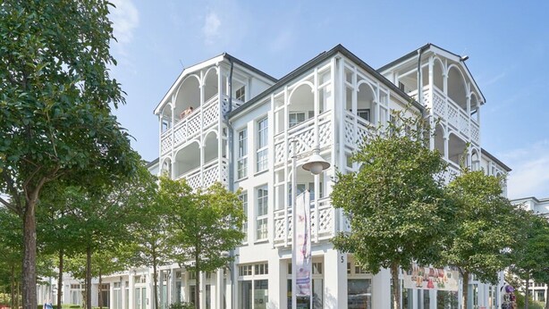 Un edificio blanco con varios balcones y un corredor cubierto, rodeado de árboles y un sendero de grava.