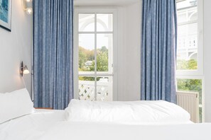 Ein Schlafzimmer mit Bett, einem Fenster mit Vorhängen, Blick auf Grünfläche und einem weißen Heizkörper.