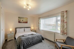 Ein Schlafzimmer mit einem Bett, Nachttischlampen, einem Heizkörper, einem Fenster mit Vorhängen, einem Sessel und einem Bild an der Wand.