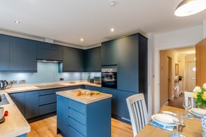 A modern kitchen with dark blue cabinets, a wooden island, and a dining area with a table set for two. - 4 Bed in Weymouth (oc-p33952) (Weymouth)
